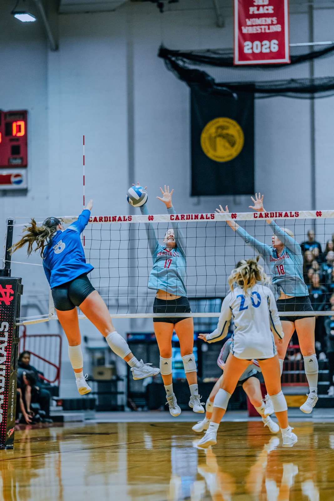 A player in blue jumps and reaches for the ball over the net as players in light blue jump to block.