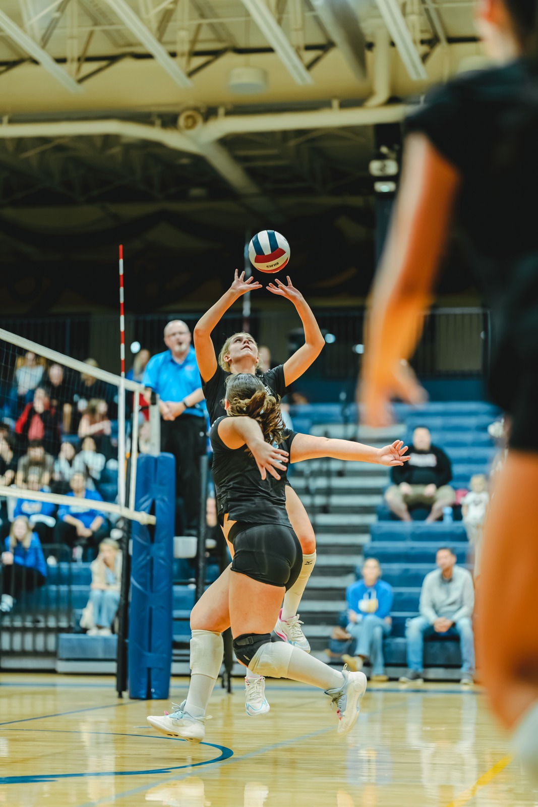 A player in black reaches overhead for the ball while another player in black crouches low.