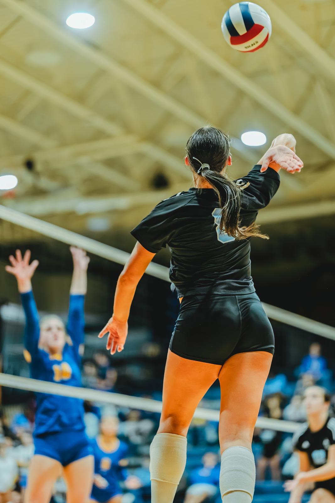 A player in black reaches for the ball over the net. A player in blue jumps with hands raised.