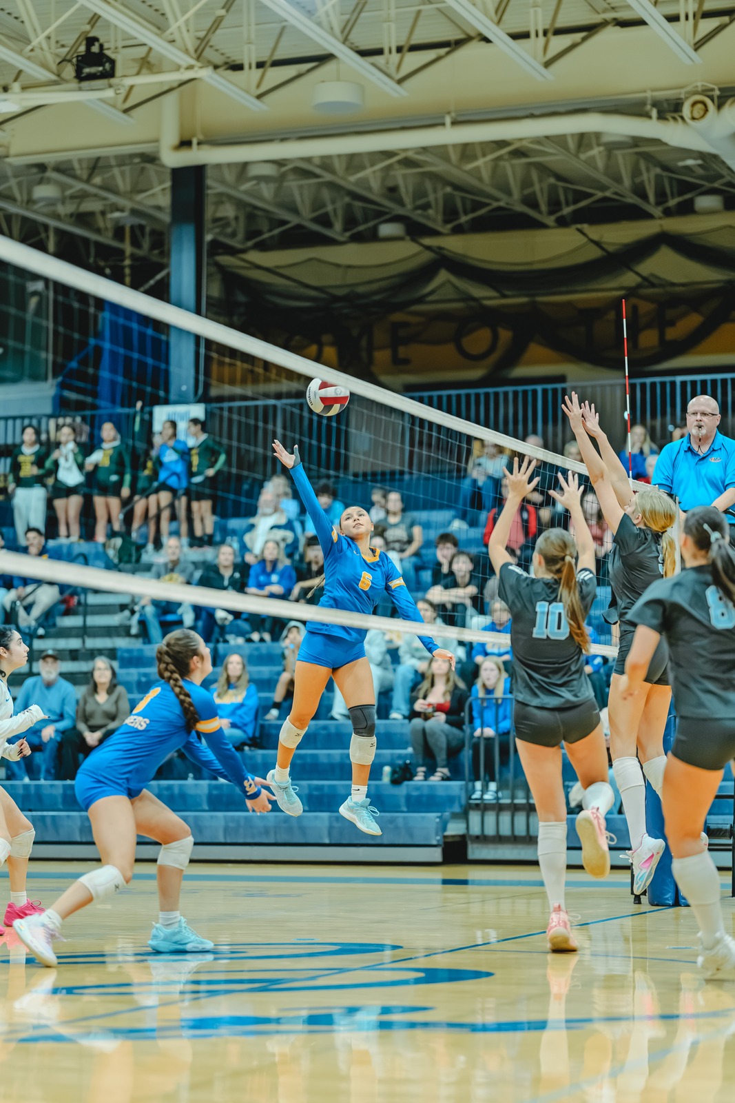 A player in blue jumps and reaches for the ball while players in gray jump to block.