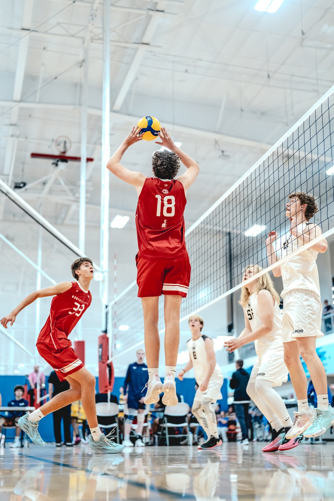 A player in a red top reaches overhead for the ball while other players in red and white uniforms are on the court.