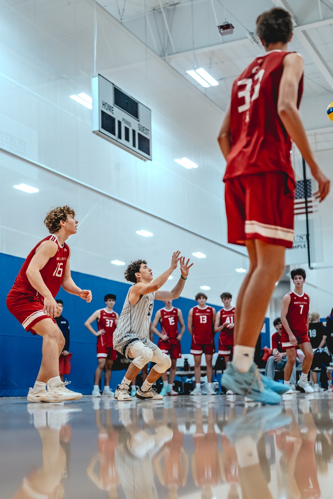 A player in a gray and white striped uniform crouches with hands extended. Players in red uniforms stand on the court.