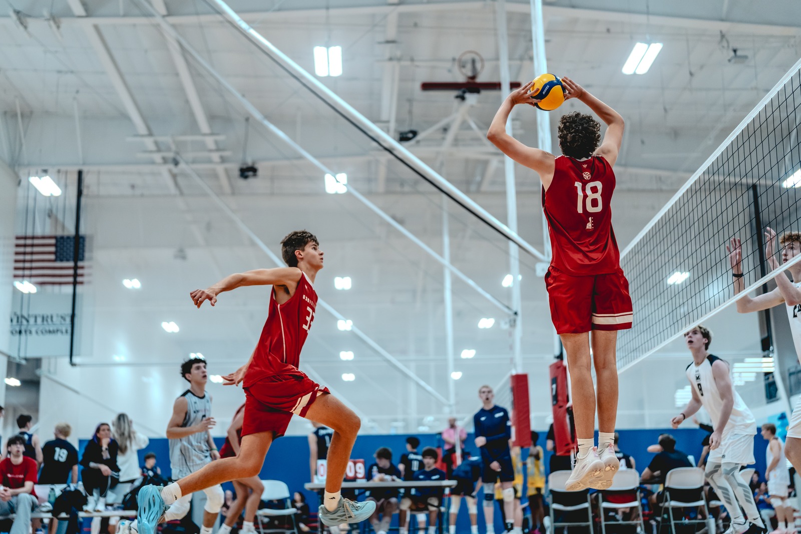 A player in red jumps with a ball overhead, while another player in red moves forward and a player in white stands ready.