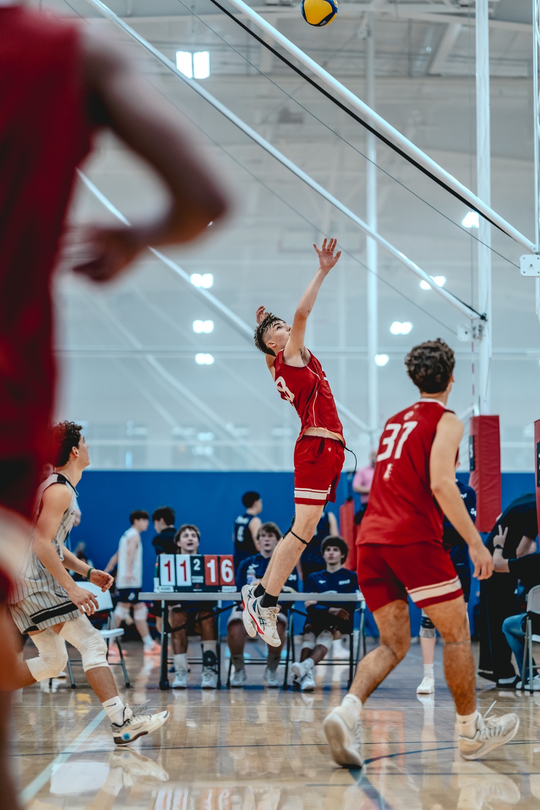 A player in red reaches overhead for the ball while jumping. Another player in red stands nearby.
