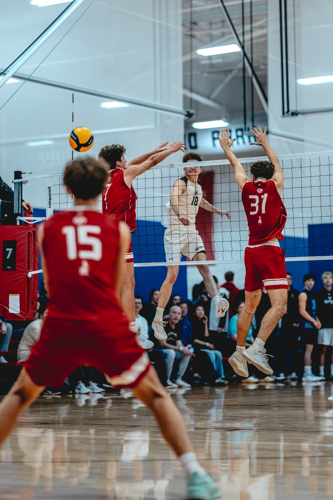 A player in red reaches up near the net as another player in white jumps and a player in red also jumps.