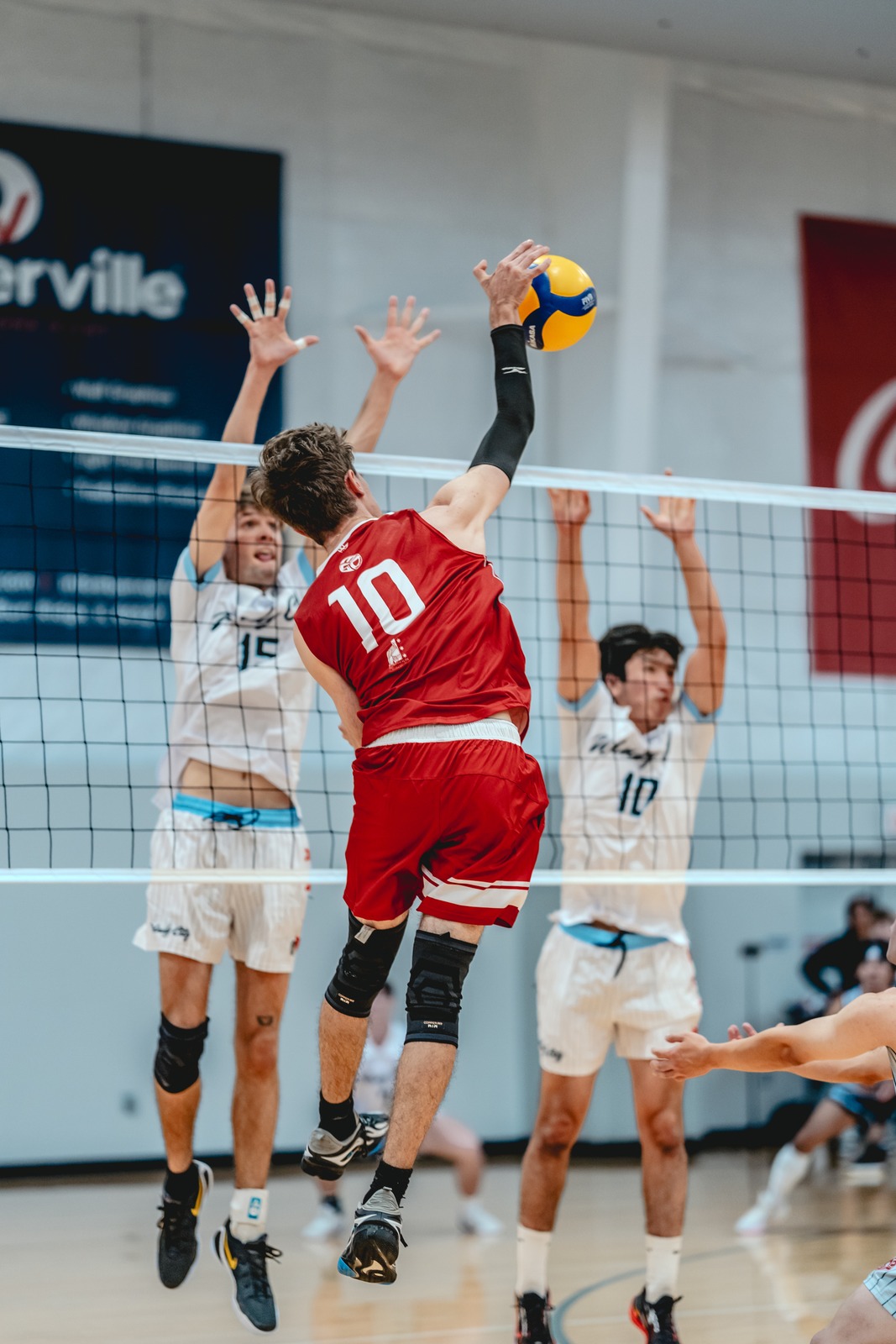 A player in red reaches overhead for the ball while players in white jump to block.