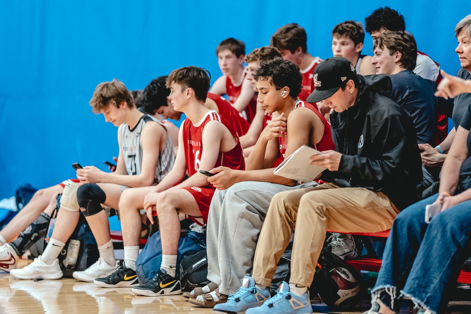 Players in red and white uniforms sit on a bench, some looking at phones, one at a notebook.